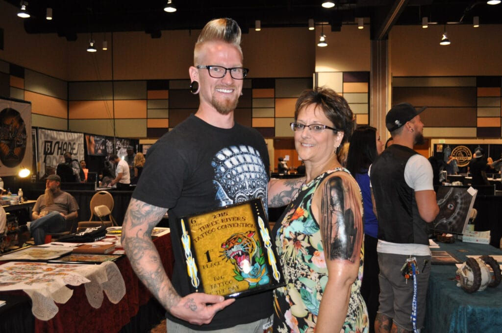 People at tattoo convention exhibit with award plaque.