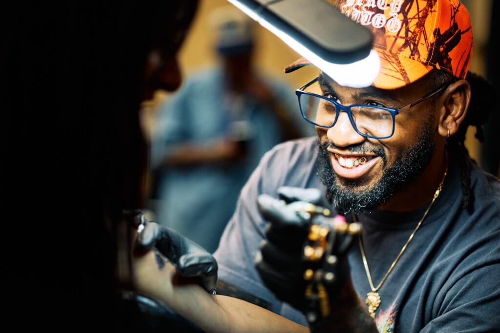 Smiling artist giving a tattoo, wearing cap and glasses.