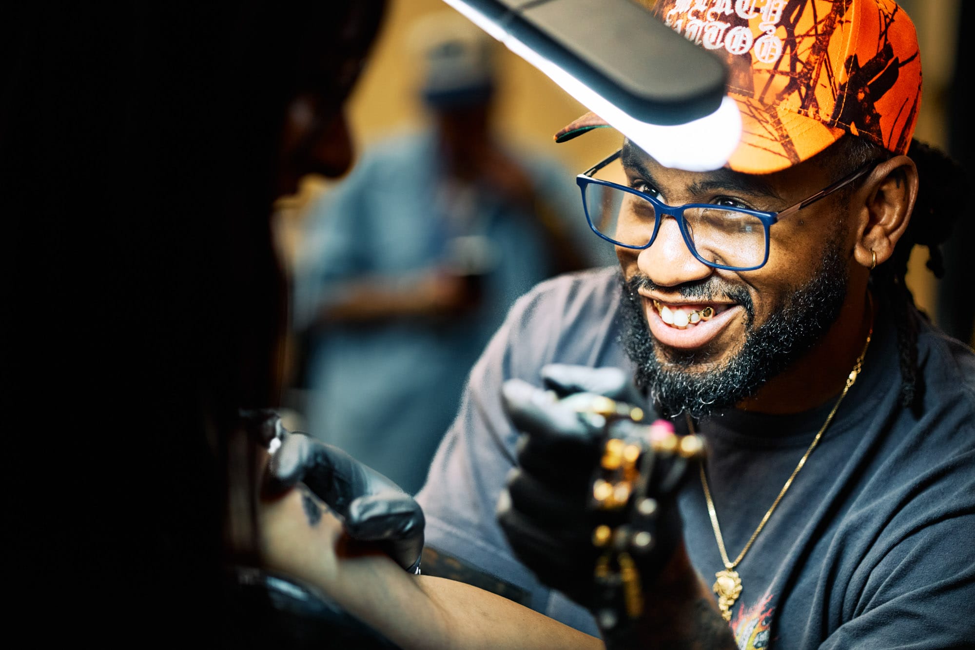 Smiling artist giving a tattoo, wearing cap and glasses.